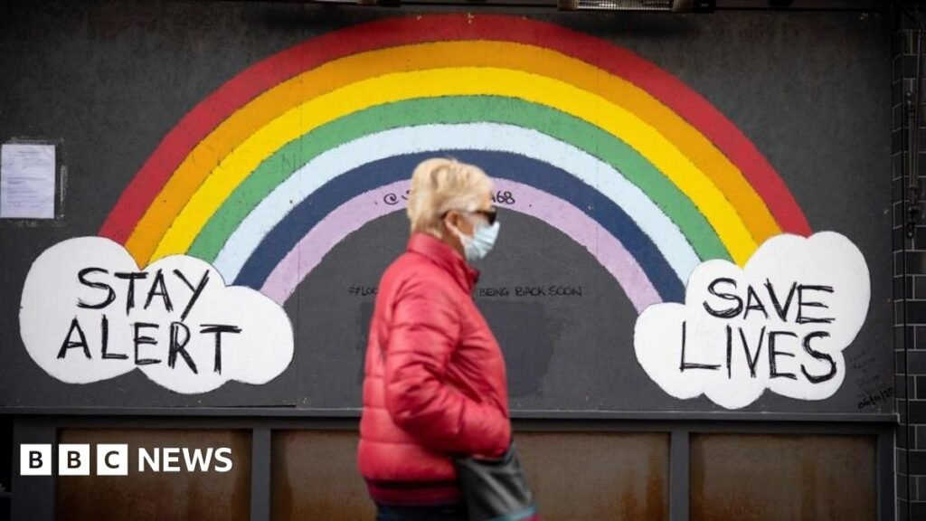 A woman wearing a face mask, sunglasses and a red coat walks past a rainbow mural on a wall which reads "Stay alert save lives".
