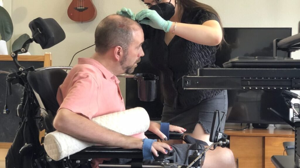 A man in a wheel chair is having a wire connected to hardware on his skull by a woman wearing medical gloves and a surgical mask.