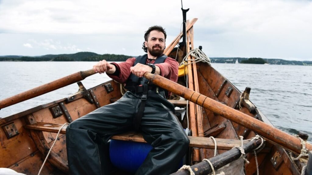 dark haired bearded man in jeans and t-shirt rowing in a viking boat