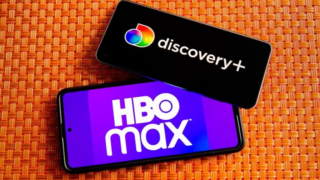 Two phones show the logos for HBO Max and Discovery Plus on an orange tablecloth
