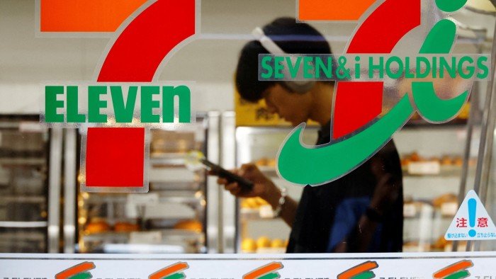 Alimentation Couche-Tard drops its $46bn pursuit of 7-Eleven owner A Tokyo 7 Eleven store, with the logo in the foreground. Seen through the glass is a man checking his phone