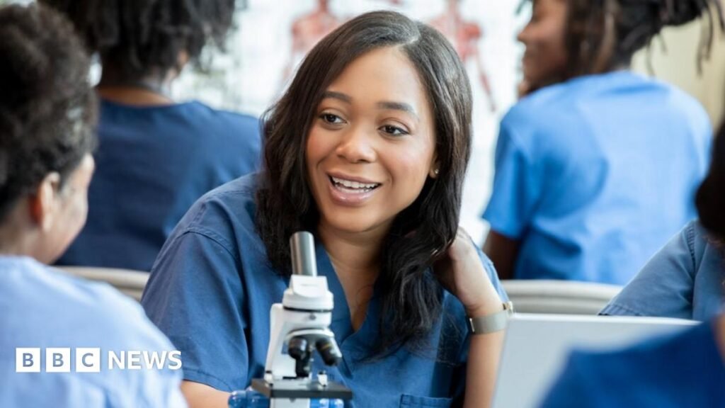 A young smiling healthcare worker is leaning on a desk with a microscope on it. She has long, dark brown hair, wears blue overalls and is surrounded by others in the same clothing.